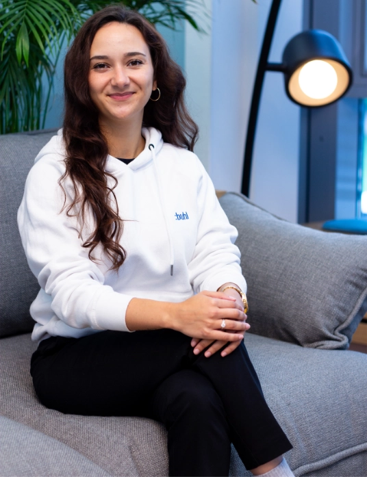 Jasmin Salchow sitzt in weißem Hoodie auf einem grauen Sofa und lächelt in die Kamera; im Hintergrund eine Stehlampe und eine grüne Pflanze