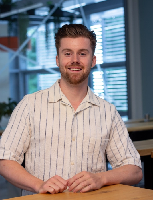 Marius Schmidt steht an einem Holztisch, trägt ein helles Hemd mit blauen Streifen und lächelt in die Kamera; im Hintergrund ein moderner Raum mit großen Fenstern
