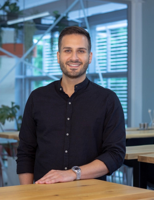 Oguz Nazli steht an einem Holztisch, trägt ein dunkles Hemd und lächelt in die Kamera, im Hintergrund ein moderner Raum mit großen Fenstern