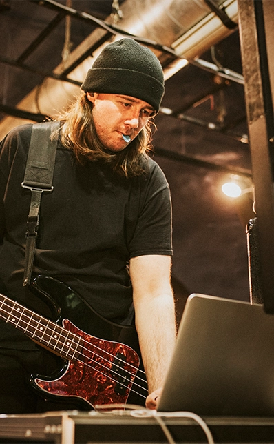 Ein Mann mit langen, dunklen Haaren und Beanie-Mütze arbeitet an einem Laptop. Um seinen Hals hängt ein E-Bass.