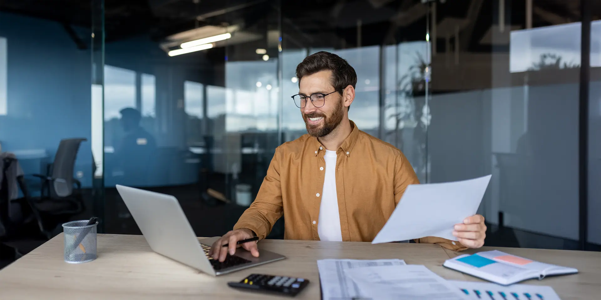 Ein junger Mann mit Brille arbeitet am einem Laptop und gleich Unterlagen ab. Auf ein Tisch steht ein Taschenrechner.