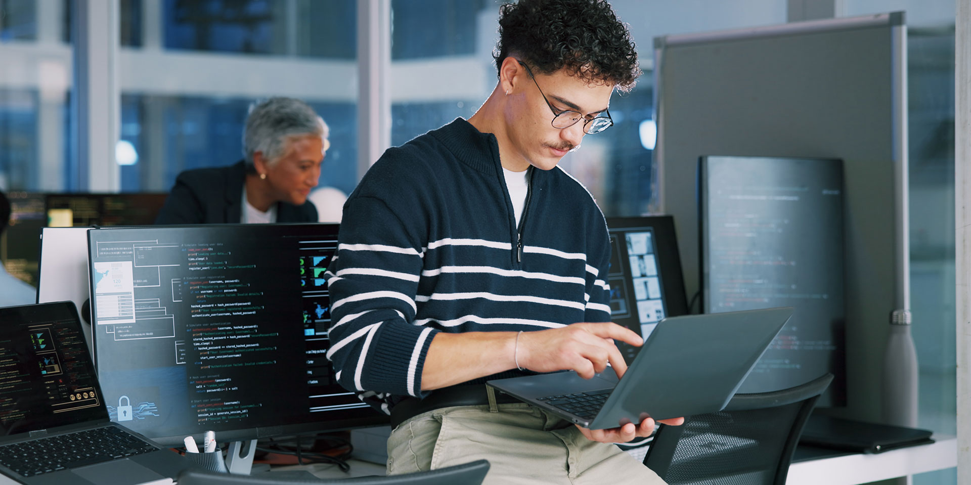 Ein junger Mann mit Brille arbeitet an einem Laptop. Im Hintergrund ein Büro mit PCs und Monitoren.