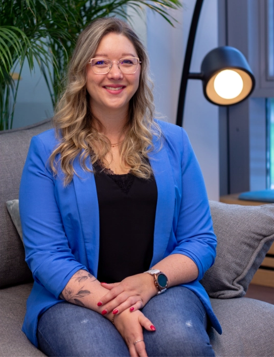 Katja Eling sitzt auf einem Sofa, trägt ein blaues Sakko, im Hintergrund eine Stehlampe und grüne Pflanze